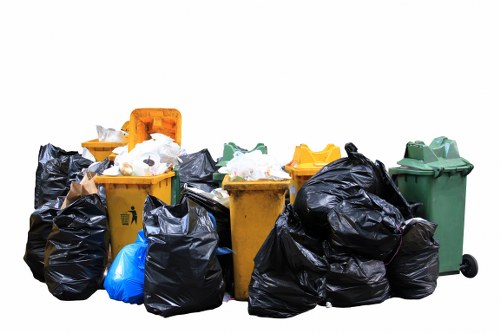 A collection of multiple waste bins and rubbish bags positioned outdoors on a paved surface, likely in a residential or commercial area within the postcode district covered by Addiscombe. The image features several yellow, green, and black wheelie bins, with some black rubbish bags cluttered around the bins, indicating a rubbish removal or clearance activity. The bins are made of durable plastic with textured surfaces and are fitted with lids, some open or slightly ajar, revealing miscellaneous waste inside. The black bags are tied at the tops and appear full, containing clutter, likely discarded household or office waste, with visible crumpled paper, plastic items, and general debris. The scene is evenly lit with natural daylight, suggesting a clear weather day. The environment surrounding the waste is plain, with no distinct background elements visible, emphasizing the focus on the refuse and bins. The image portrays typical rubbish collection objects, aligning with the services provided by Office Clearance Addiscombe in the local London area, aiding industrial or domestic rubbish disposal needs.