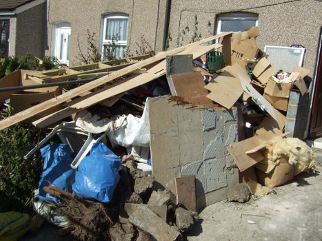 A large pile of mixed household waste and construction debris is situated on a residential street pavement, with a background of multi-storey buildings and greenery. The pile includes broken concrete slabs, wooden planks, cardboard boxes, and various discarded items such as packaging and fabric. The materials display different textures and colors, notably grey concrete, light brown wood, and brown corrugated cardboard, with some items partially covered by a white sheet or tarpaulin. The debris is stacked chaotically, occupying most of the visible sidewalk area near the curb, with some pieces leaning against each other and others scattered around. The scene suggests an accumulation of rubbish awaiting collection or clearance, consistent with services offered by Office Clearance Addiscombe in the Addiscombe area. The lighting indicates a bright, sunny day, highlighting the variety of textures and cluttered arrangement in the waste pile, typical of waste removal scenarios in local residential or commercial contexts.