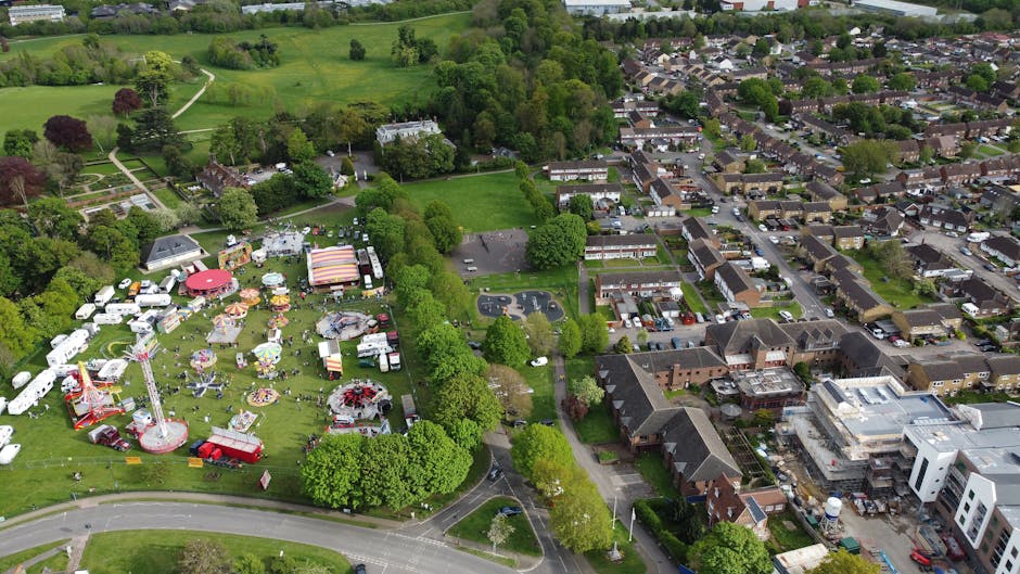 An aerial view of a fairground situated within a park, featuring several colourful amusement rides, including a red and white striped carousel and a tall tower ride, surrounded by lush green trees and