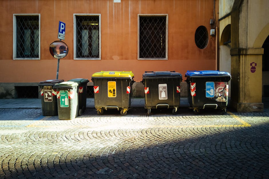 A collection of various plastic containers and jerry cans arranged outdoors, including a large blue recycling bin with a white label, a yellow jerry can with a black cap, a red fuel canister, a blue s