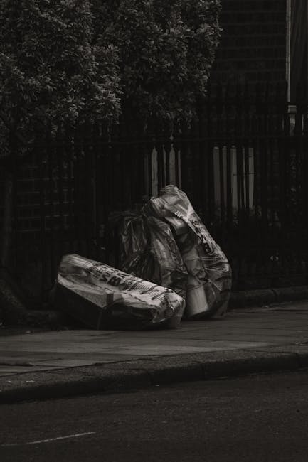 Two large black rubbish bags filled with waste rest on a pavement in front of a dark metal fence, with dense foliage visible behind the fence. The bags appear to be made of heavy-duty plastic and are 