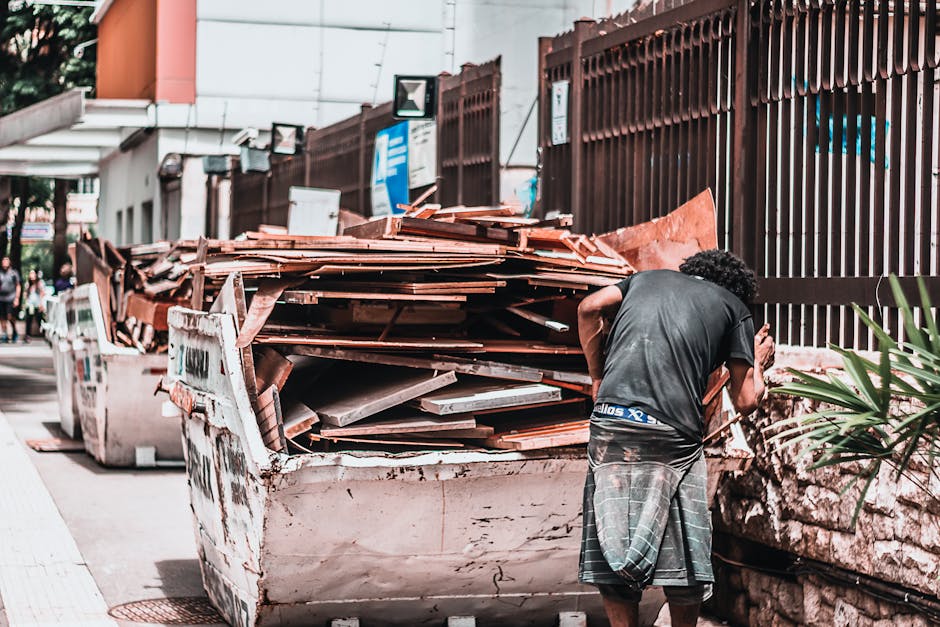 A small dark red flatbed truck is parked on a city street in front of a modern building with large windows and a concrete facade. The truck's cargo area is filled with various types of rubbish, includ