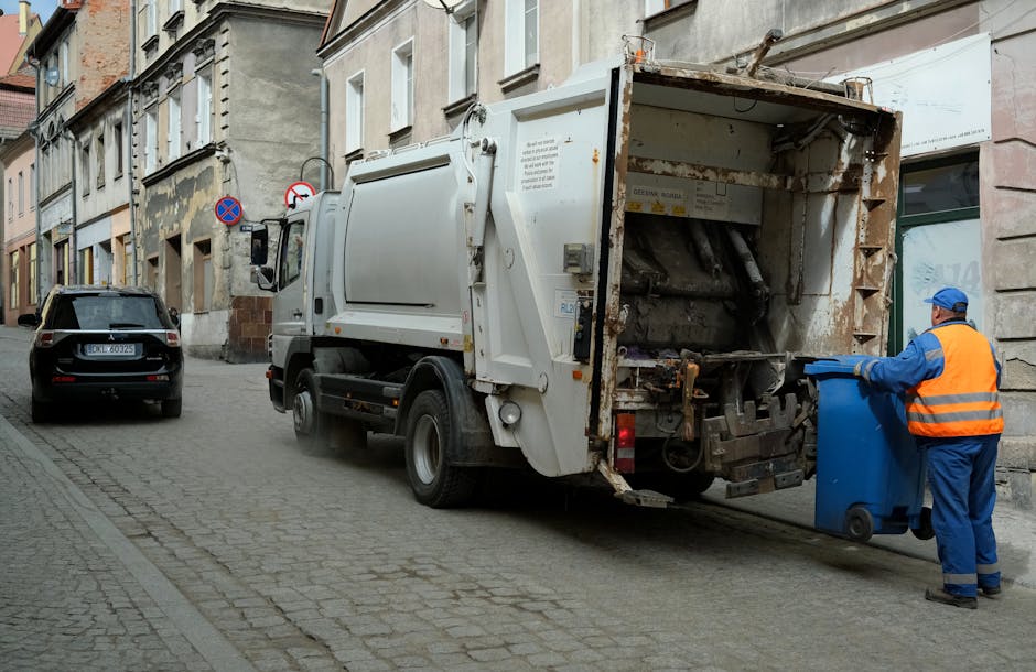 A street scene with a white waste collection truck positioned parallel to the curb, actively engaged in rubbish removal on an urban street lined with multi-story buildings featuring stone and concrete